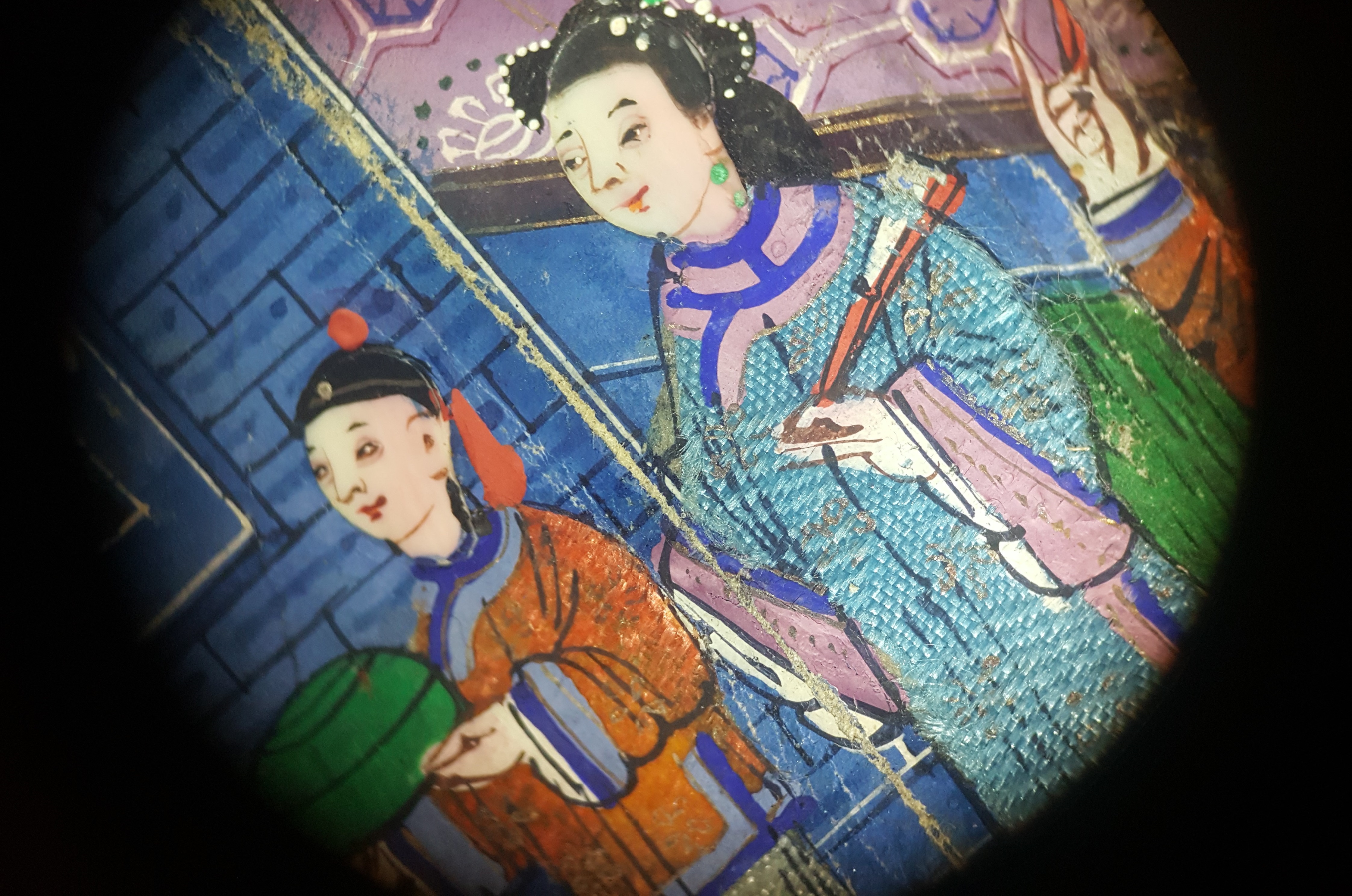 A close detail of two small figures on the fan, of a young man in an orange robe and a woman in a blue robe in front of a blue wall, showing the finely painted woven textile and ivory faces